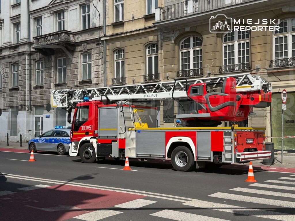 Czerwony wóz strażacki z podnośnikiem i niebieski radiowóz policji zatrzymane na ulicy, w tle zabytkowy budynek.