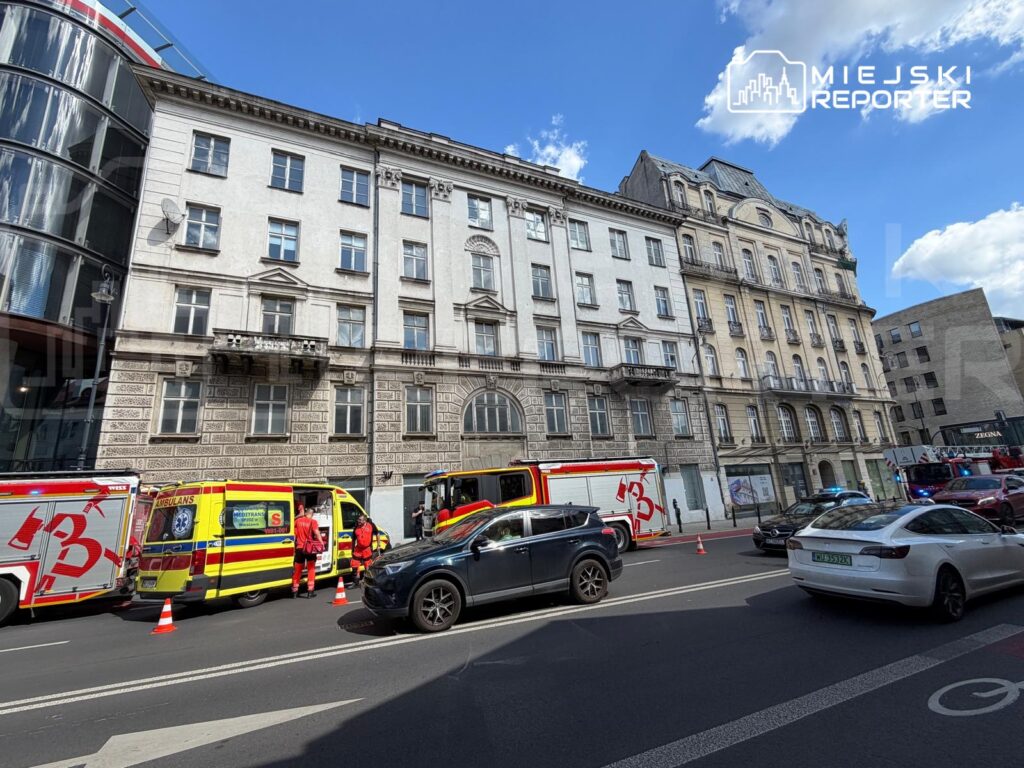 Ratownicy medyczni w czerwonych strojach pracują przy ambulansach na ulicy, obok zabytkowego budynku.