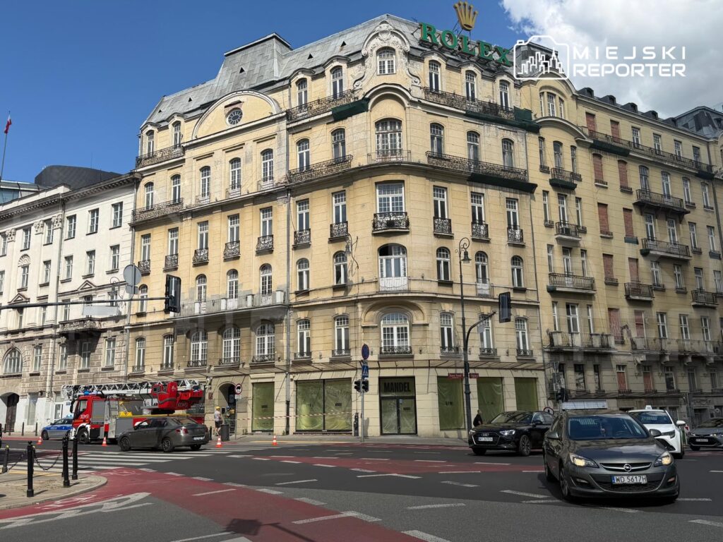 Zabytkowy budynek z żółtą elewacją i detalami architektonicznymi, z widocznym znakiem Rolex oraz wozem strażackim na ulicy.