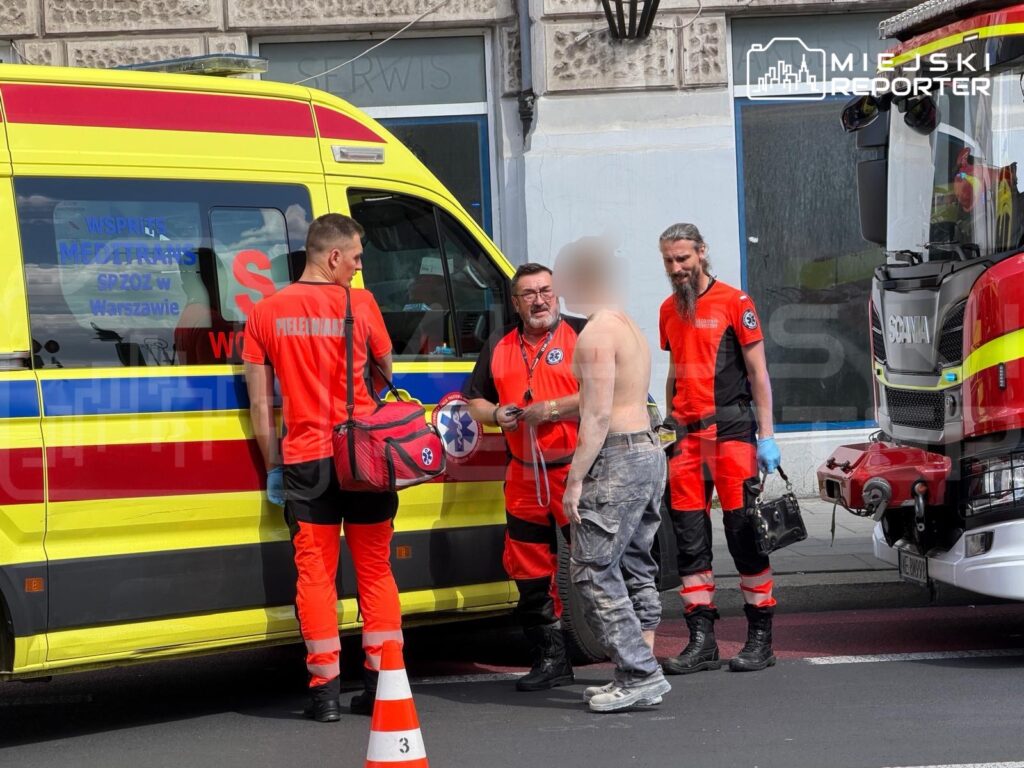 Czterech ratowników w pomarańczowych strojach rozmawia z mężczyzną bez koszuli obok karetki i wozu strażackiego.
