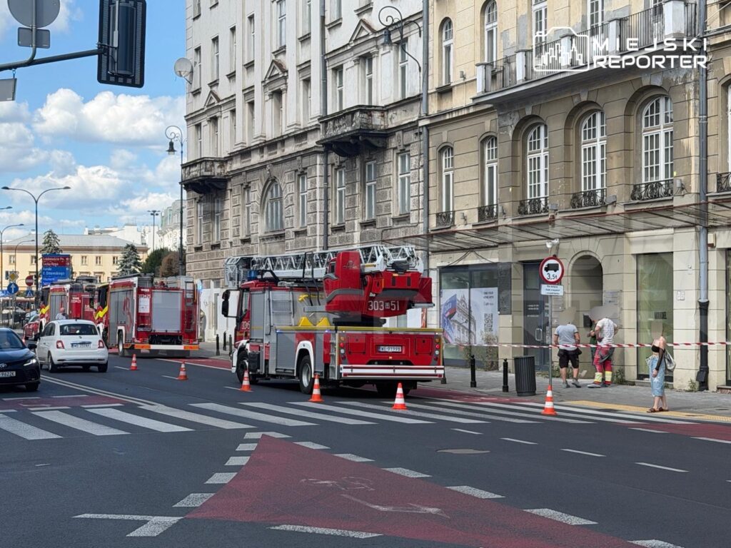 W ulicy stoi woz strażacki z drabiną, a obok dwóch mężczyzn pracuje przy budynku, wyznaczono strefę zabezpieczoną taśmą.