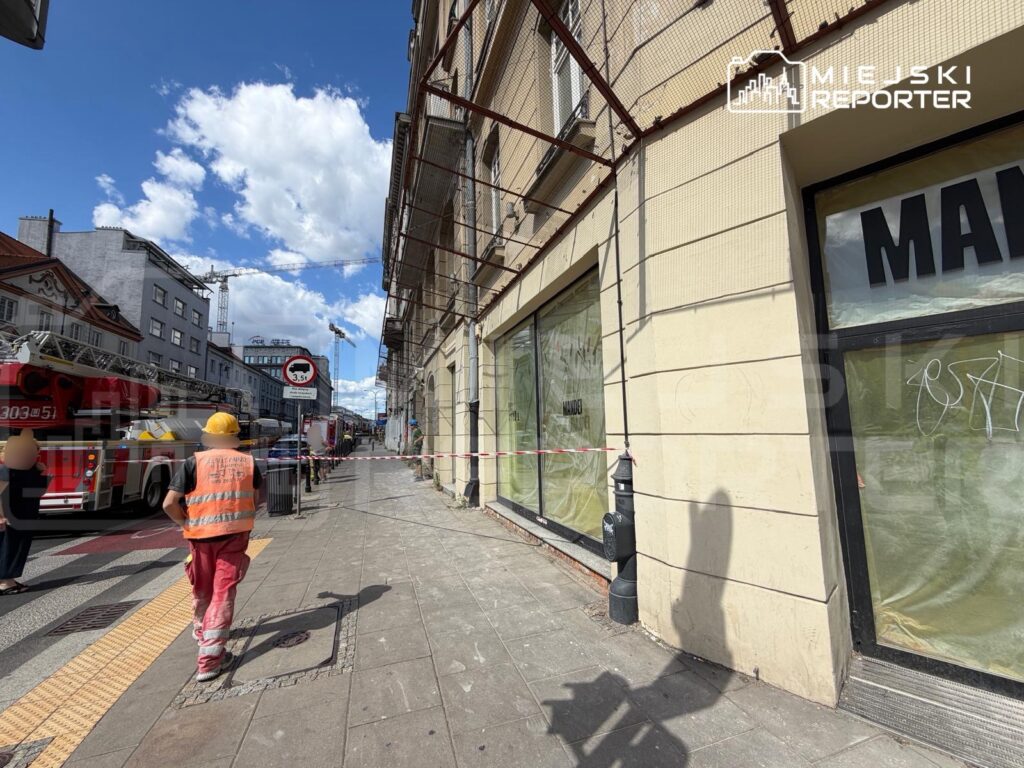 Robotnik w pomarańczowej kamizelce kieruje się w stronę budynku z zasłoniętymi oknami, obok parkują wóz strażacki i blokada ulicy.