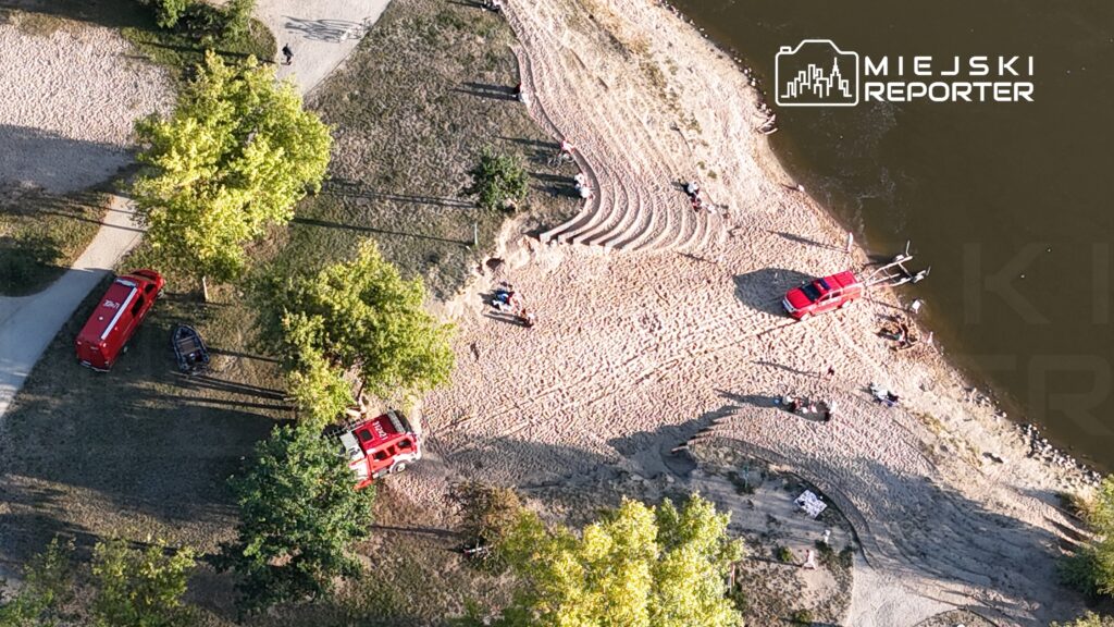 Czerwony wóz strażacki i łódź na plaży, obok ludzi relaksujących się na piasku nad brzegiem wody.
