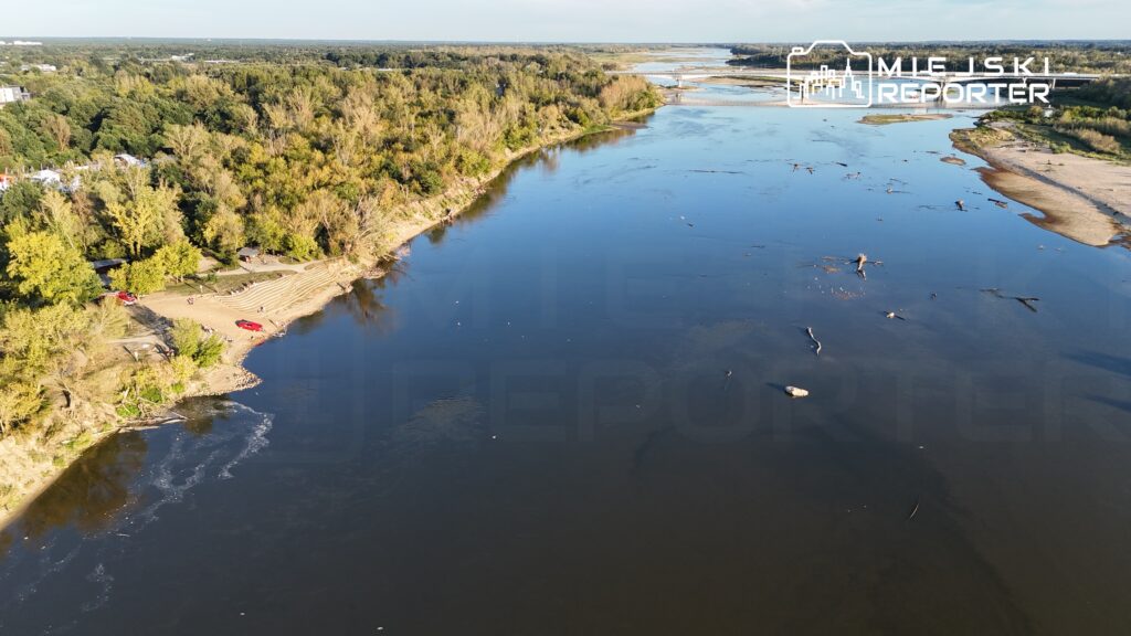 Rzeka płynie wśród zielonych drzew, a na plaży widać ludzi i dwa czerwone samochody zaparkowane na piasku.