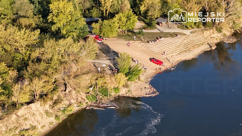 Plażowicze relaksują się na piaszczystym brzegu rzeki, obok dwóch czerwonych wozów strażackich.