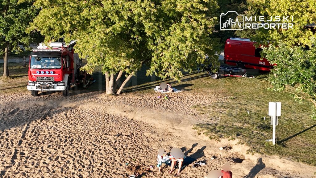 Czerwony wóz strażacki stoi na plaży, obok wypoczywające osoby na kocach, w tle drzewa i piasek.
