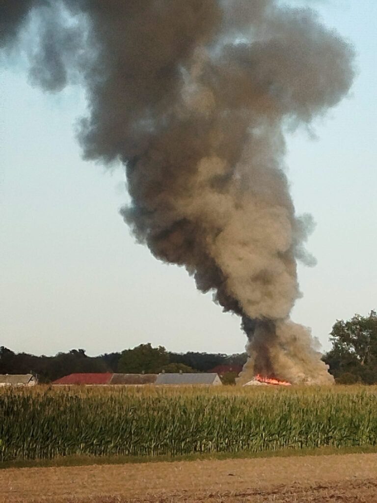 Wielka chmura dymu unosząca się nad polem, w tle widać płonący budynek oraz sąsiednie zabudowania.