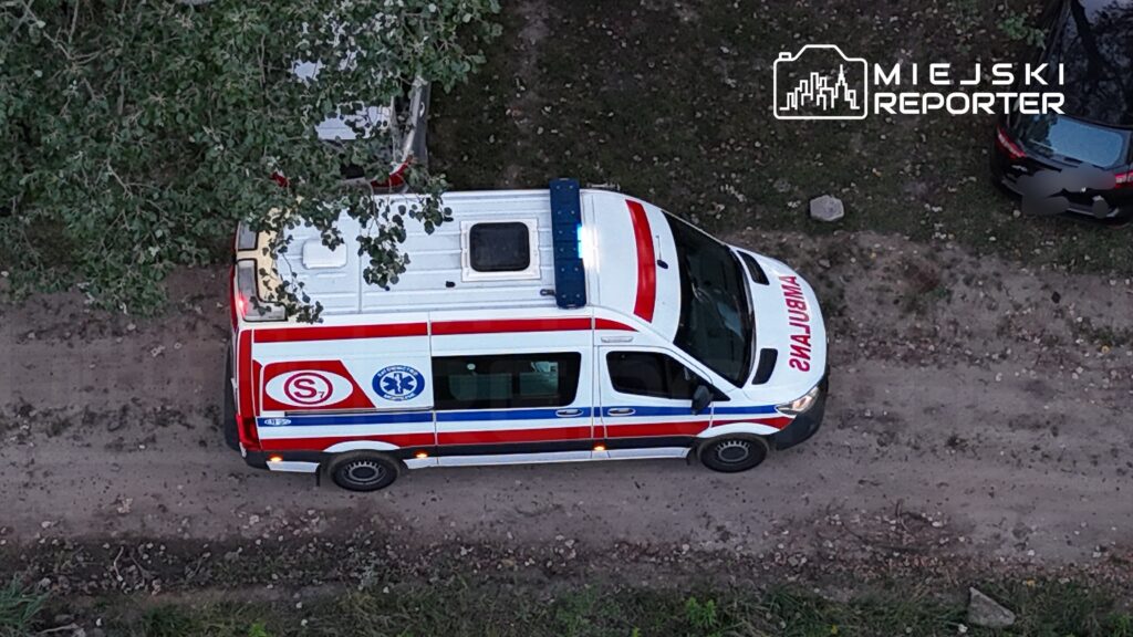 Ambulans z niebieskimi światłami zaparkowany na leśnej drodze, otoczony drzewami i innymi pojazdami.