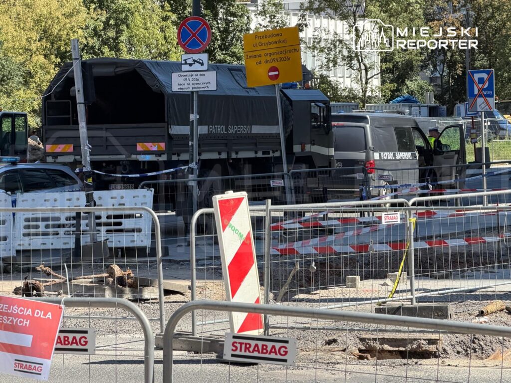 Samochód patrolowy oraz ciężarówka z napisem "PATROL SAPERSKI" na ruchliwej ulicy, otoczone barierkami budowlanymi.