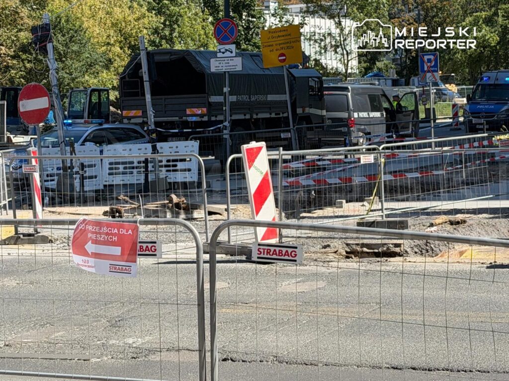 Na ulicy widać policyjny samochód oraz wojskowy pojazd, otoczone barierkami budowlanymi. Znajduje się tam znak "DO PRZEJŚCIA DLA PIESZYCH".