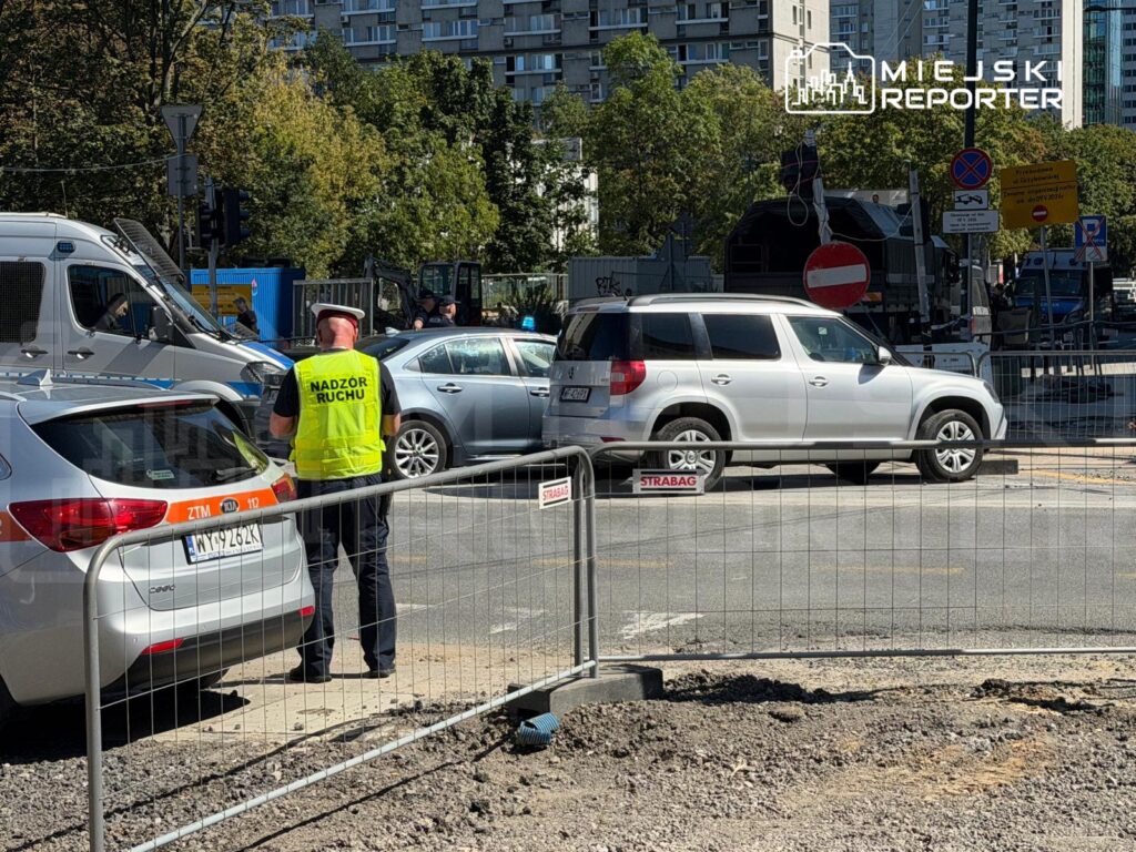 Mężczyzna w kamizelce odblaskowej z napisem "Nadzór Ruchu" kontroluje ruch drogowy na zablokowanej ulicy.