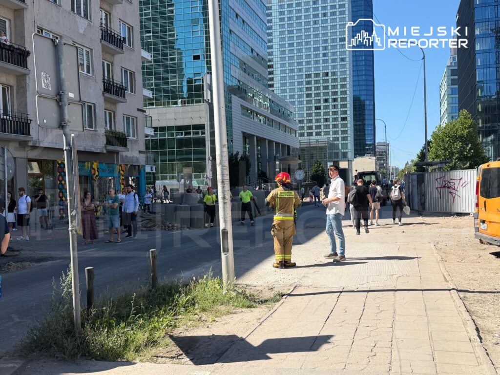 Grupa ludzi obserwująca strażaka w mundurze, który rozmawia z mężczyzną w białej koszuli na ulicy w centrum miasta.