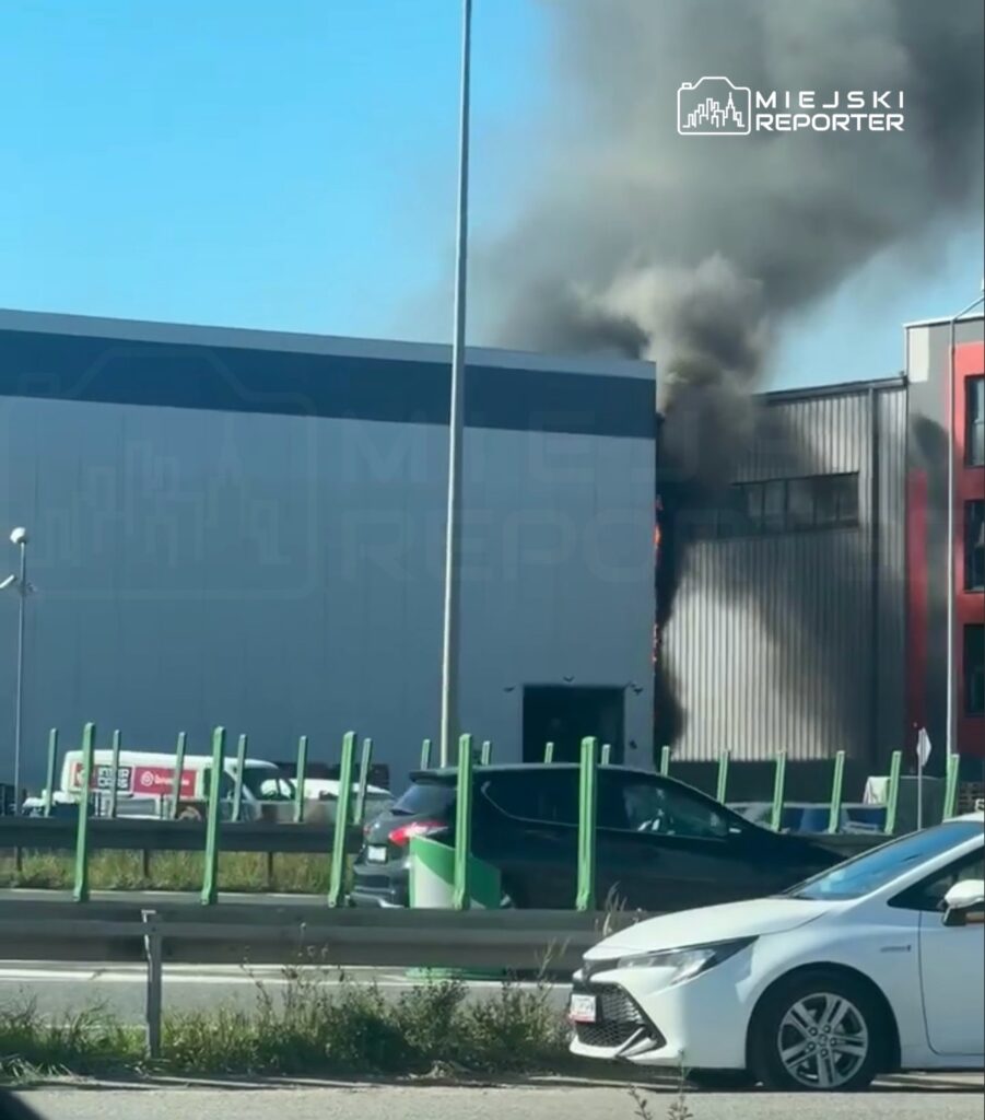 Czarny dym wydobywający się z budynku przemysłowego przy drodze, w tle samochody na ruchliwej trasie.