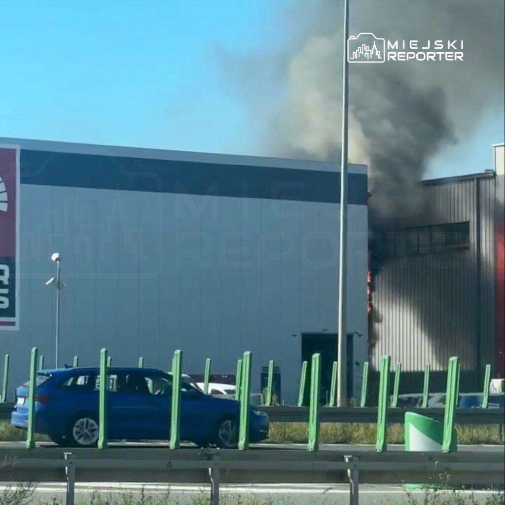 Na tle niebieskiego nieba wydobywa się czarny dym z budynku przemysłowego, obok przejeżdża niebieski samochód osobowy.