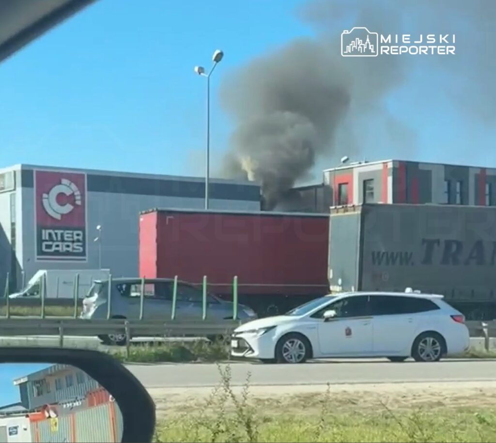 Czarny dym unosi się z budynku z logo "Inter Cars" w otoczeniu zaparkowanych samochodów na drodze.