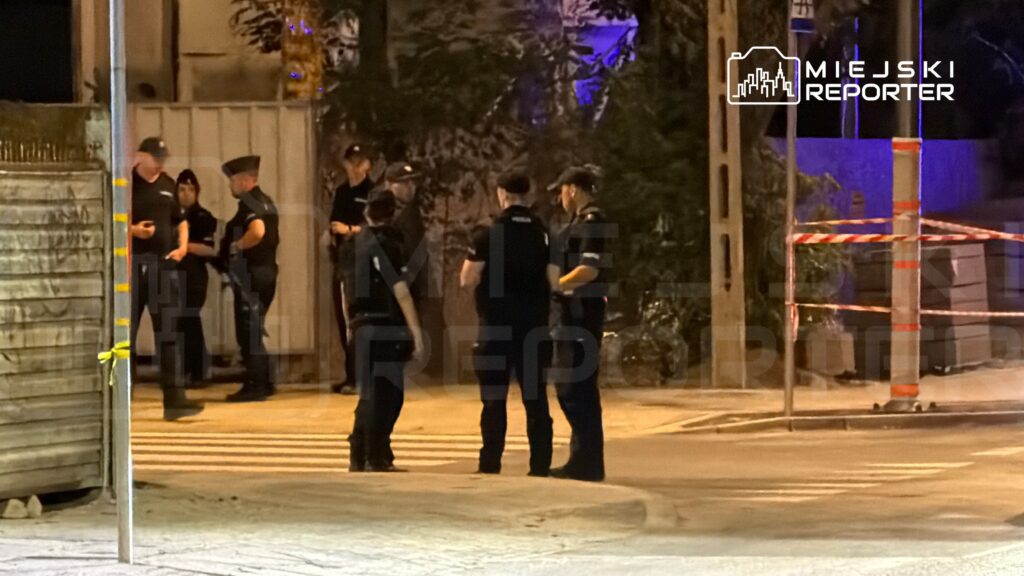 Grupa policjantów w mundurach rozmawia na ulicy, w tle widać oświetlone drzewo i zamknięty teren.