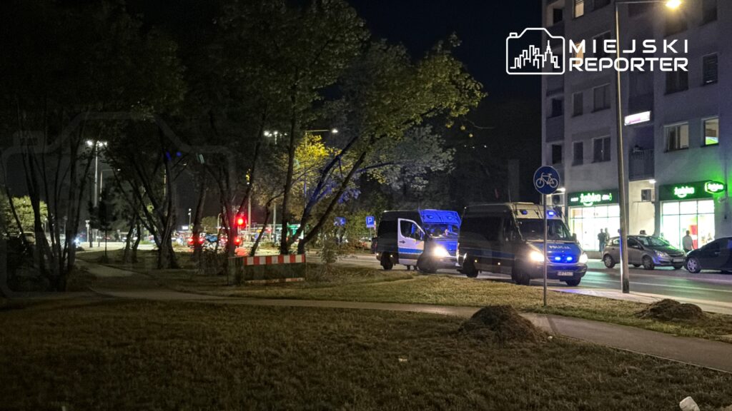 Policyjne vany zatrzymane na ulicy w nocy, z włączonymi sygnałami świetlnymi, w pobliżu sklepów.
