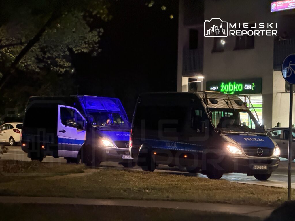Dwa radiowozy policyjne z włączonymi światłami na parkingu w nocy, w tle widoczny sklep spożywczy.