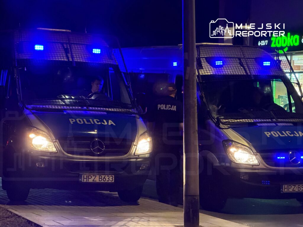 Policyjny pojazd z niebieskimi światłami stoi na ulicy, a policjanci rozmawiają obok. W tle widać sklep.