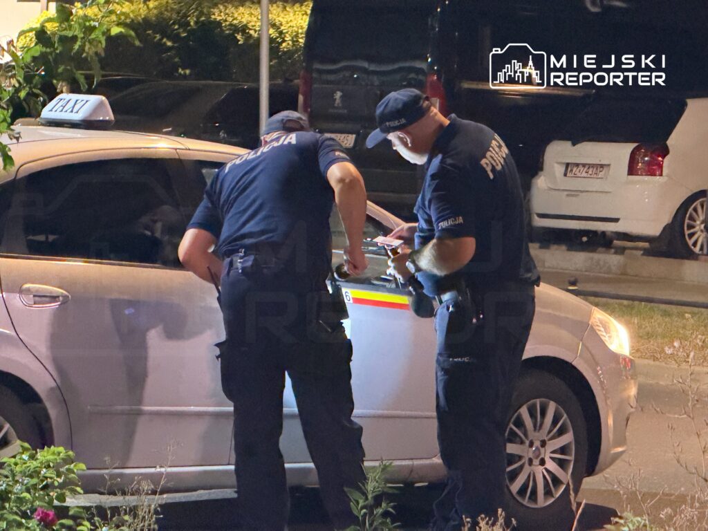 Dwóch policjantów sprawdza dokumenty kierowcy taksówki na nocnej ulicy.