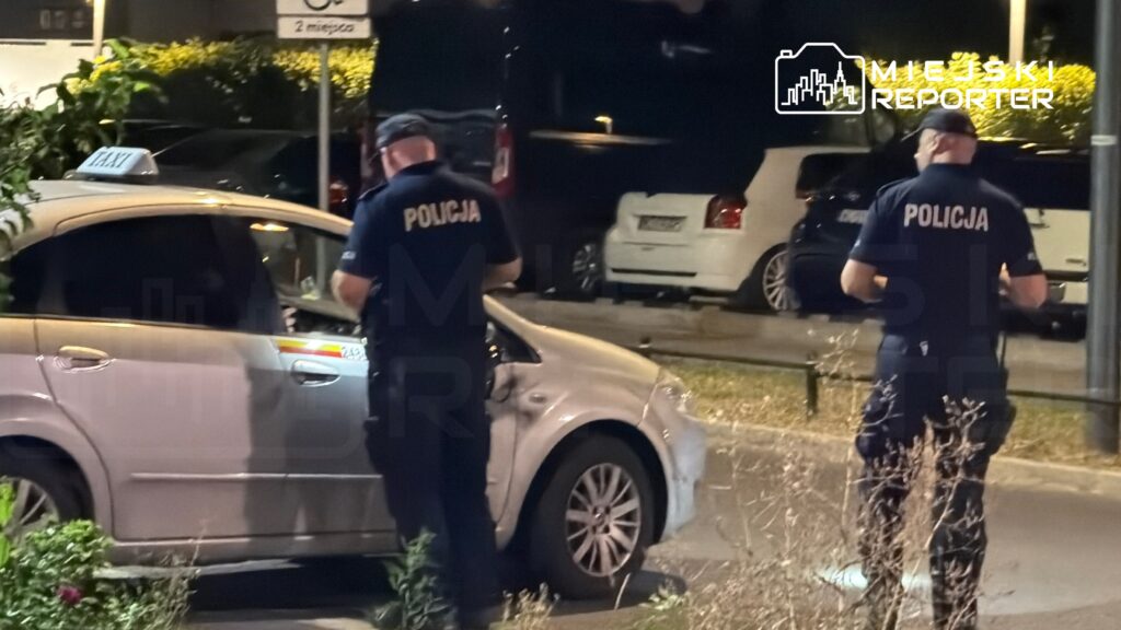 Dwaj policjanci kontrolują taksówkę zaparkowaną na ulicy w nocy.