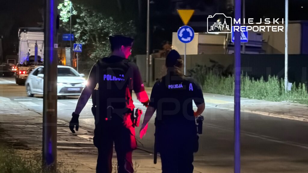 Dwóch policjantów w mundurach patroluje ulicę nocą, obok przejeżdżają samochody.