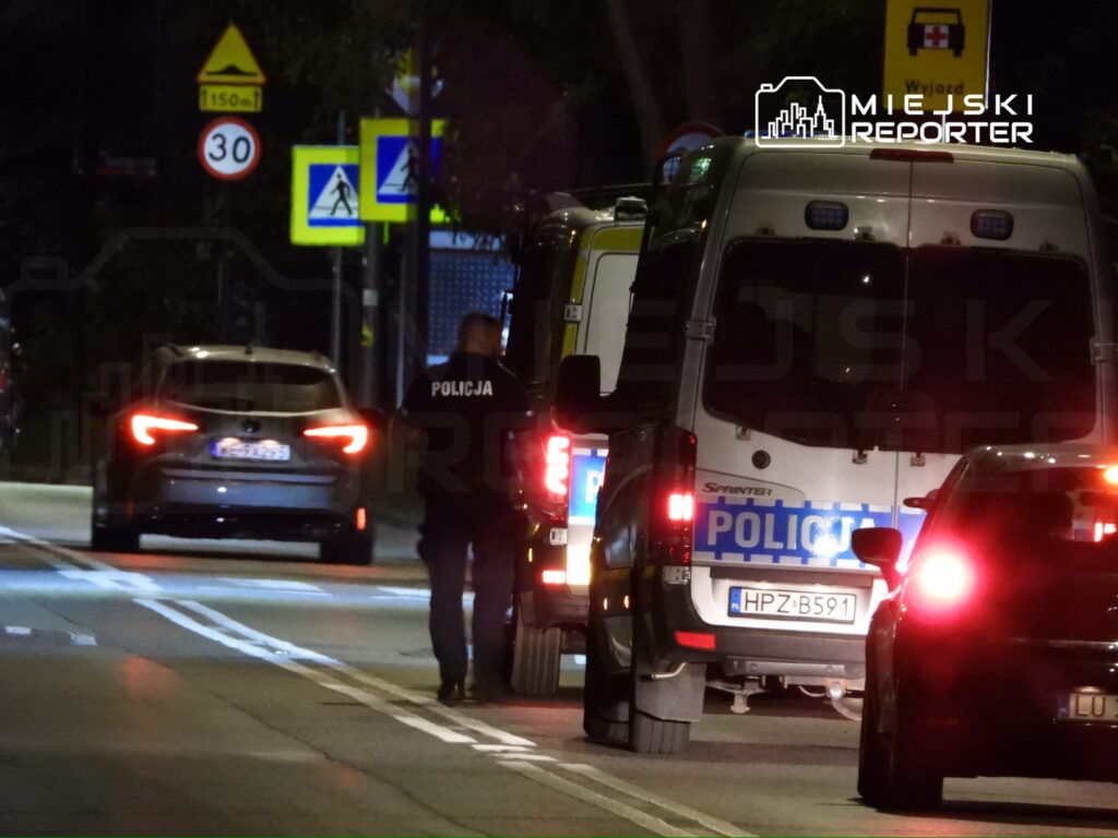 Policjant w mundurze sprawdzający sytuację obok radiowozów na oświetlonej ulicy nocą.