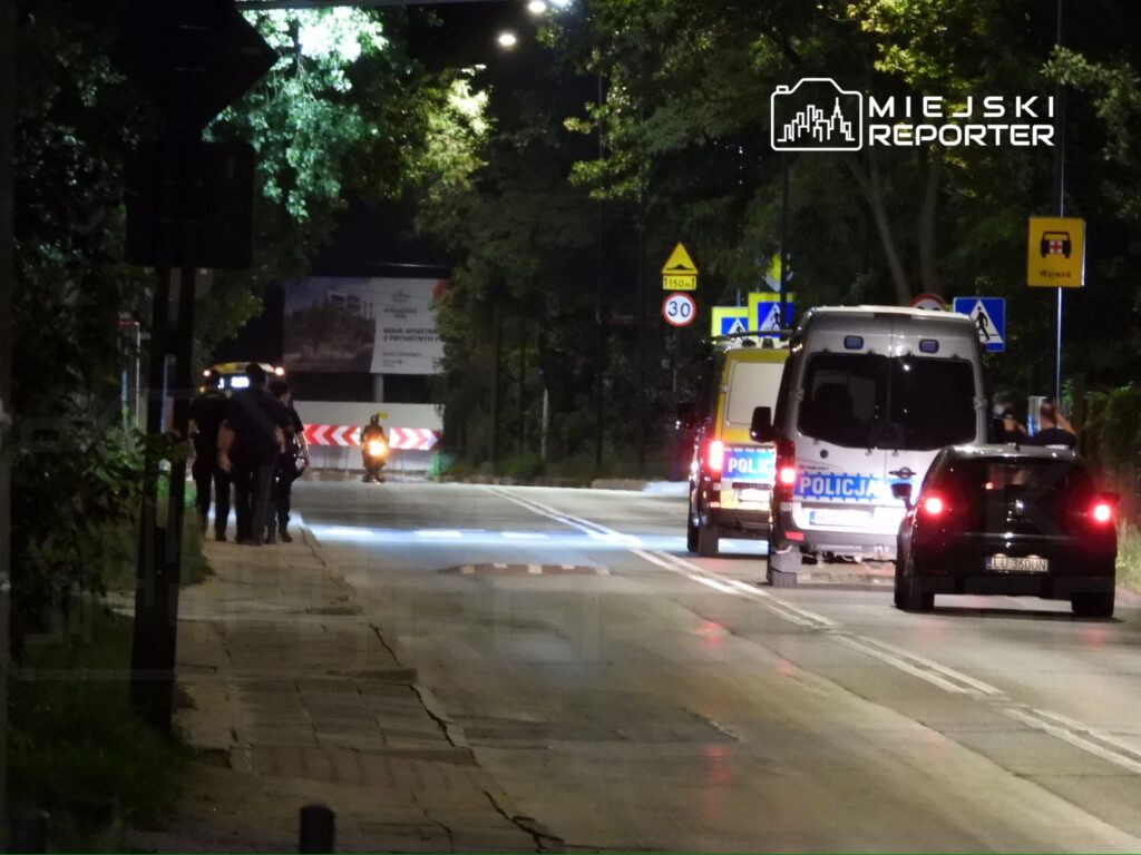 Grupa policjantów idzie chodnikiem w nocy w pobliżu zaparkowanych radiowozów, z oświetleniem ulicznym w tle.