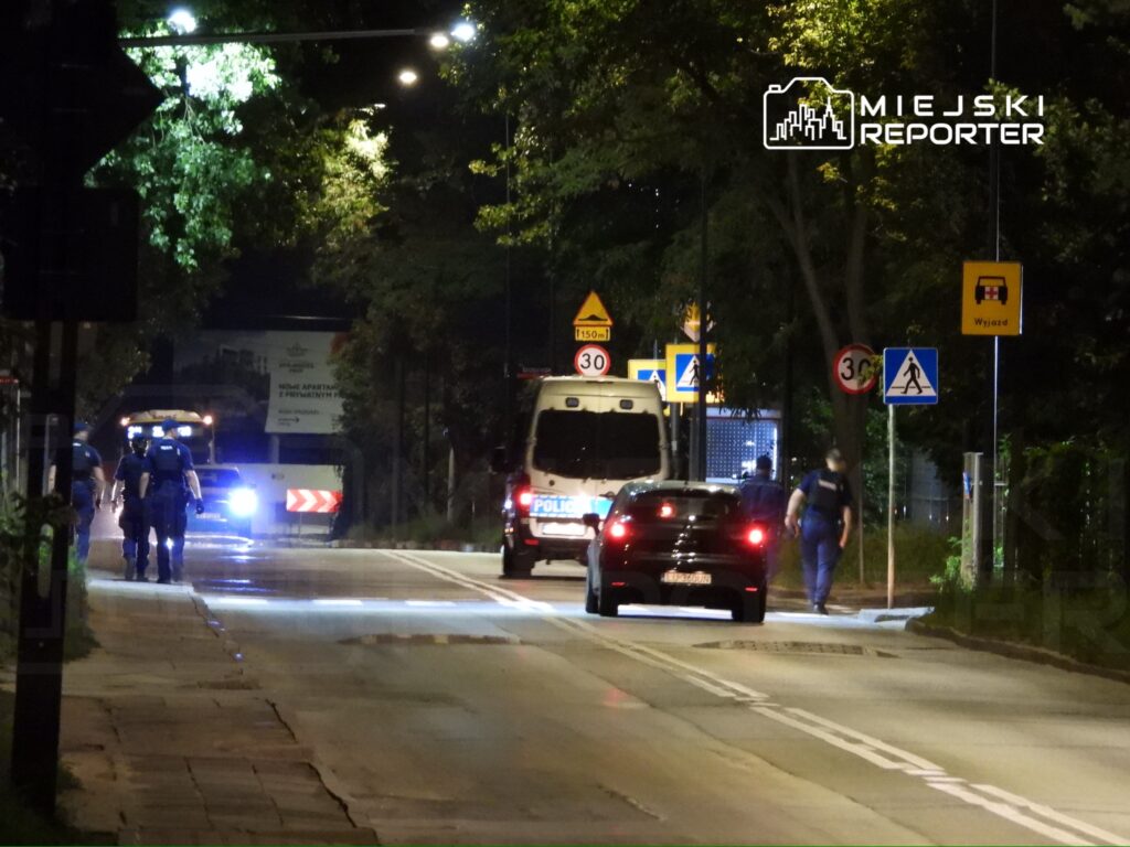 Grupa policjantów patroluje oświetloną ulicę nocą, obok stoją policyjne pojazdy.