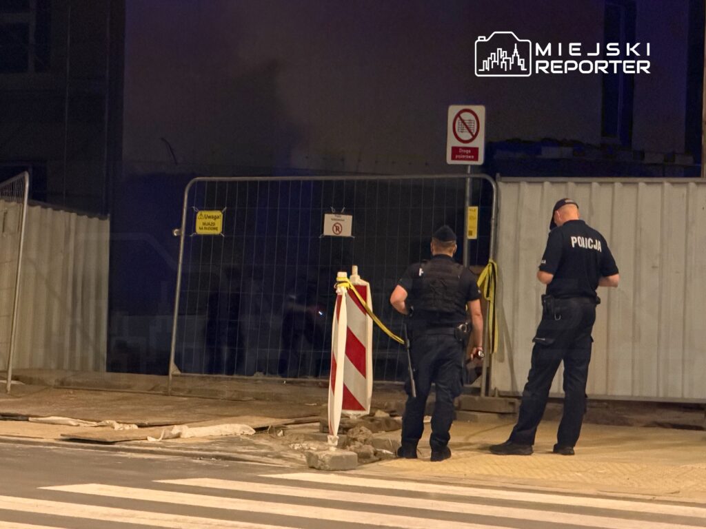 Dwóch policjantów w mundurach stoi przy ogrodzeniu budowy, sprawdzając teren w nocy.