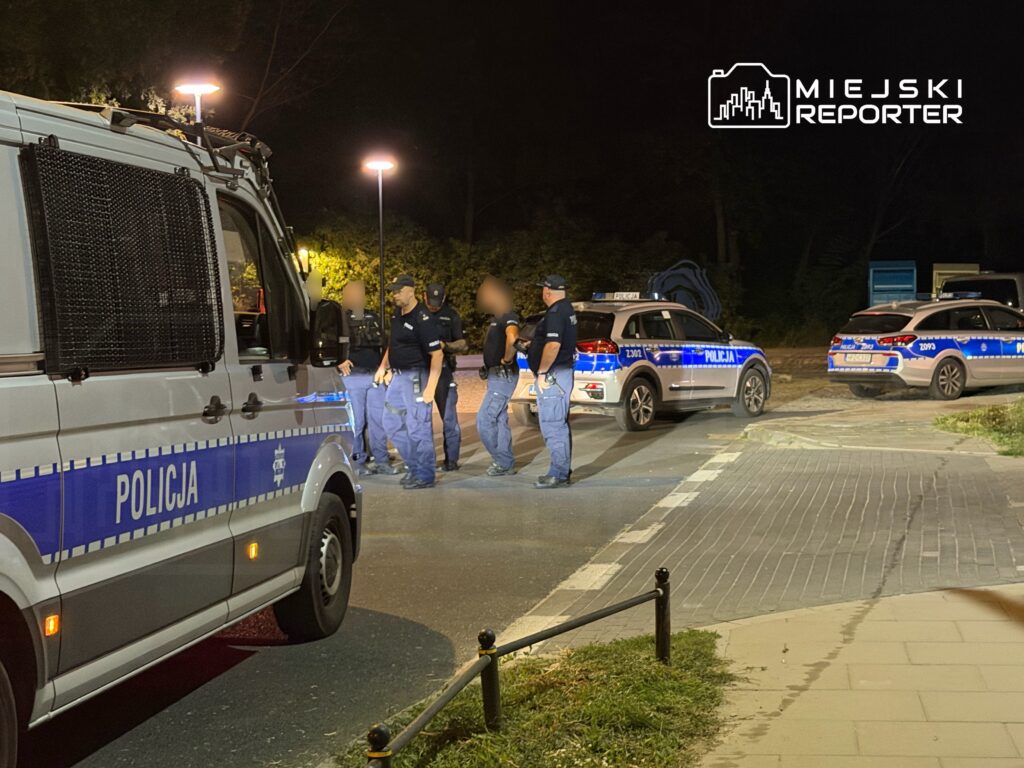 Grupa policjantów w mundurach rozmawia obok radiowozów na oświetlonym parkingu w nocy.