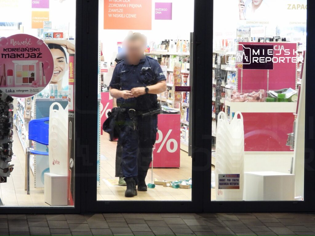 Policjant w mundurze przemieszcza się wewnątrz drogerii, na tle półek z kosmetykami i promocjami.