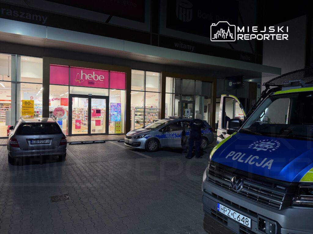 Policjant w mundurze rozmawia z innym funkcjonariuszem przy radiowozie przed apteką.