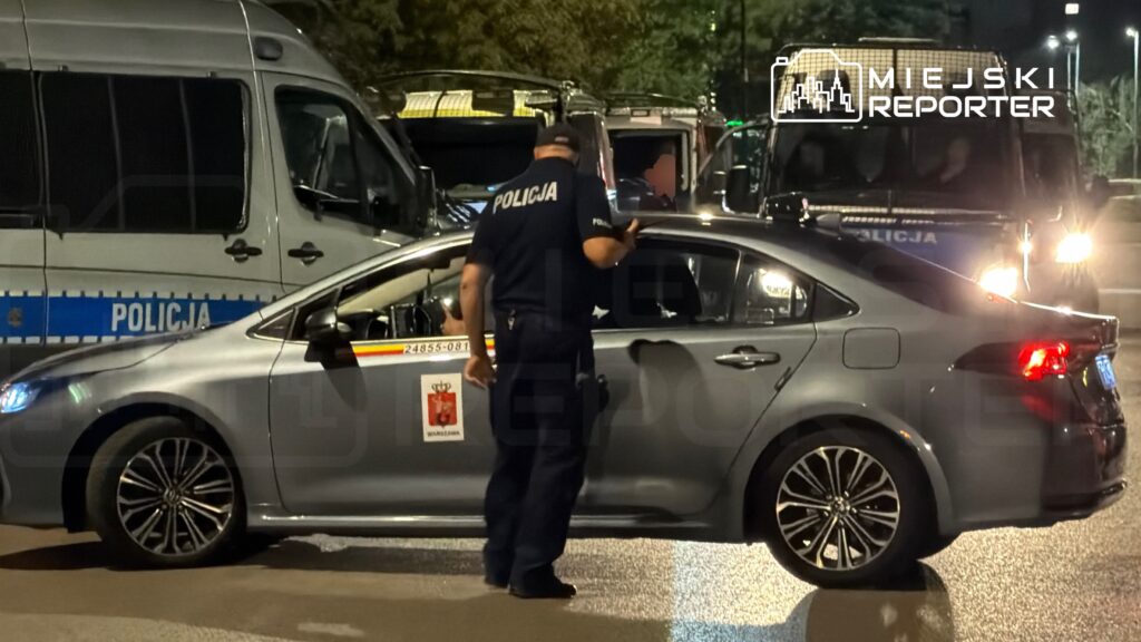 Policjant kontrolujący pasażera w szarym samochodzie osobowym podczas nocnej akcji w mieście.