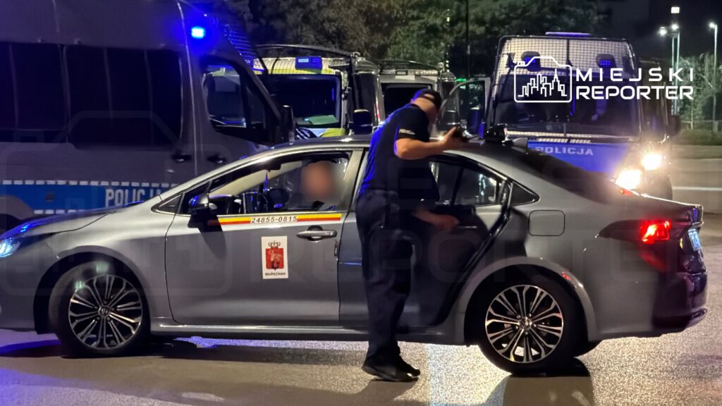 Policjant sprawdzający wnętrze radiowozu na tle zaparkowanych pojazdów policyjnych w nocy.