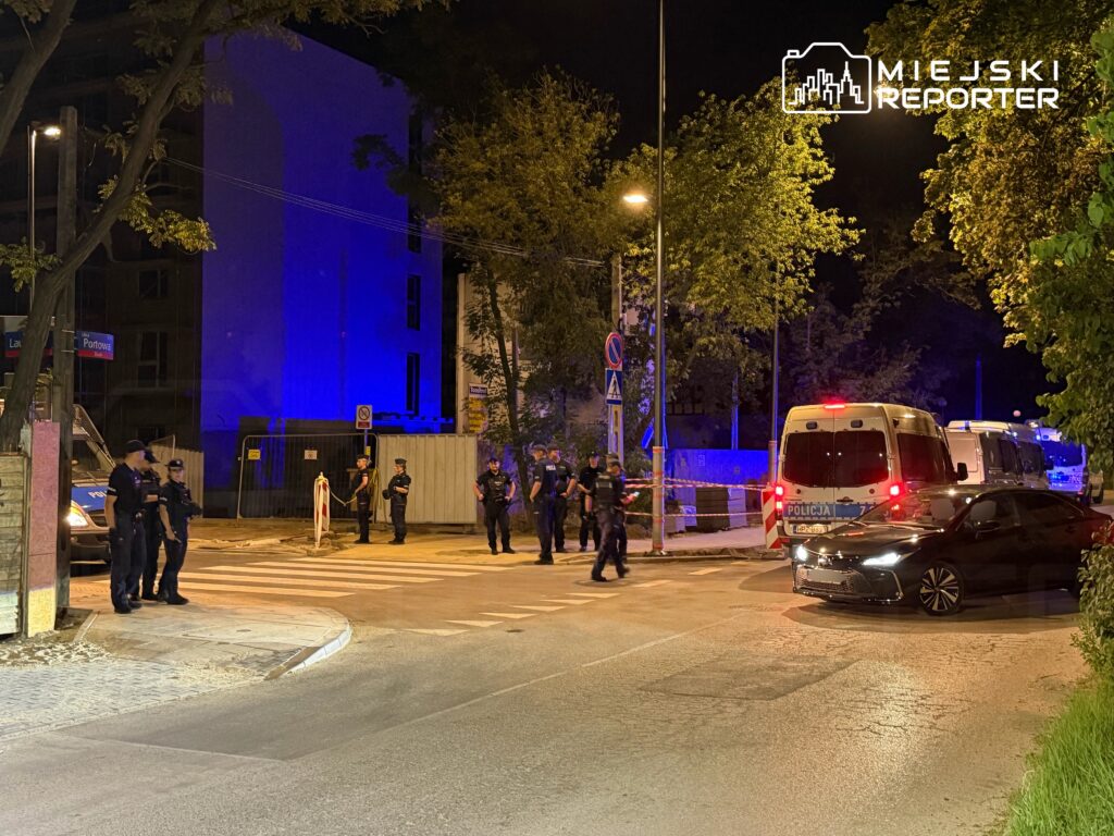 Grupa policjantów w mundurach zabezpiecza miejsce zdarzenia nocą, obok zaparkowane radiowozy i samochód osobowy.