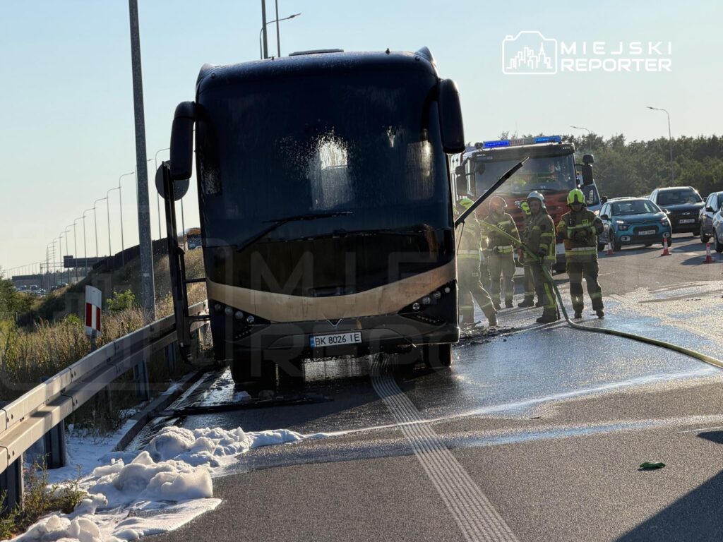 Ogień w autobusie gaszony przez strażaków na poboczu drogi, w tle przejeżdżające samochody.
