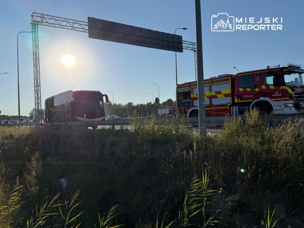 Autobus stoi na poboczu drogi, obok znajduje się wóz strażacki z włączonymi sygnałami świetlnymi. Słońce świeci na tle nieba.