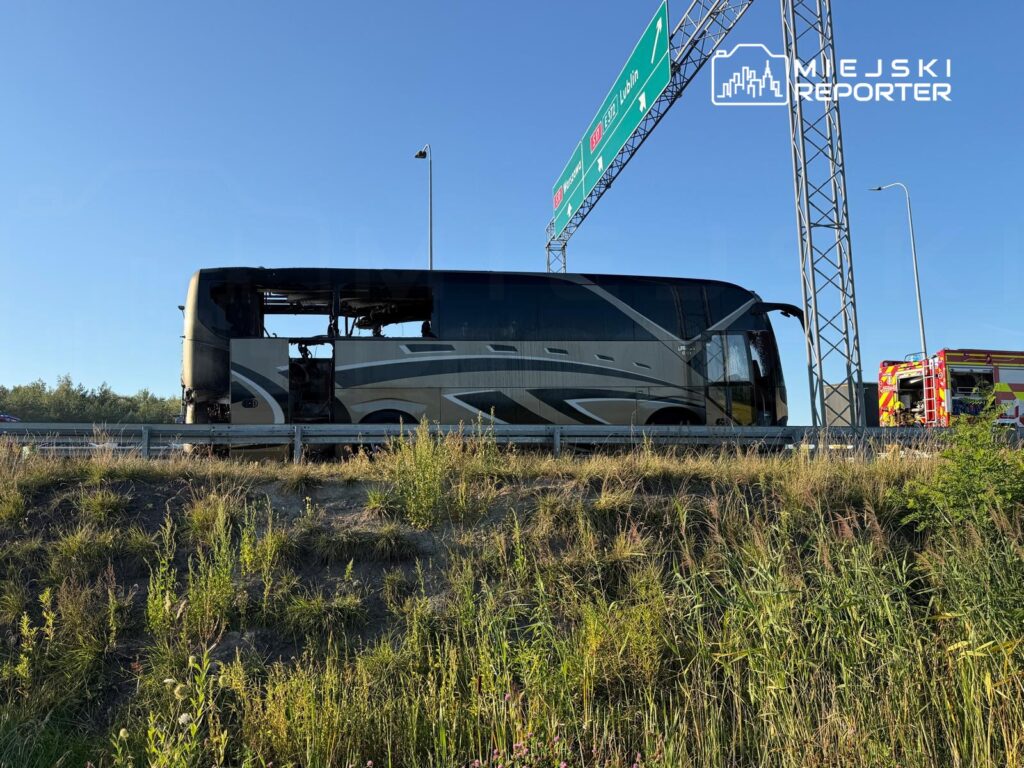 Spalony autobus stojący przy drodze, obok wozu strażackiego, na tle nieba i zarośli.