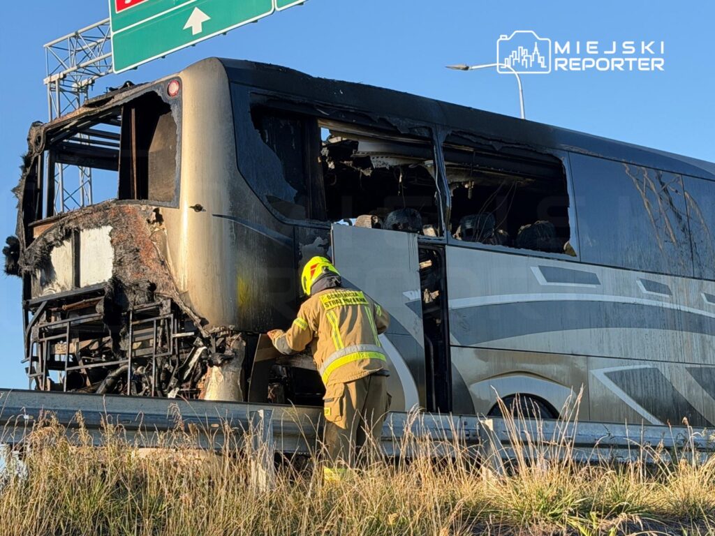 Strażak bada uszkodzony autobus z popalonym nadwoziem na poboczu drogi, w tle znaki drogowe.