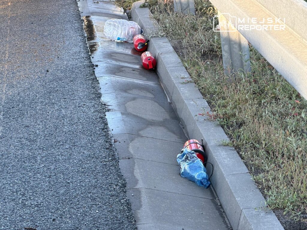 Na poboczu drogi leżą plastikowe butelki i opakowania po napojach, obok woda spływa wzdłuż krawężnika.