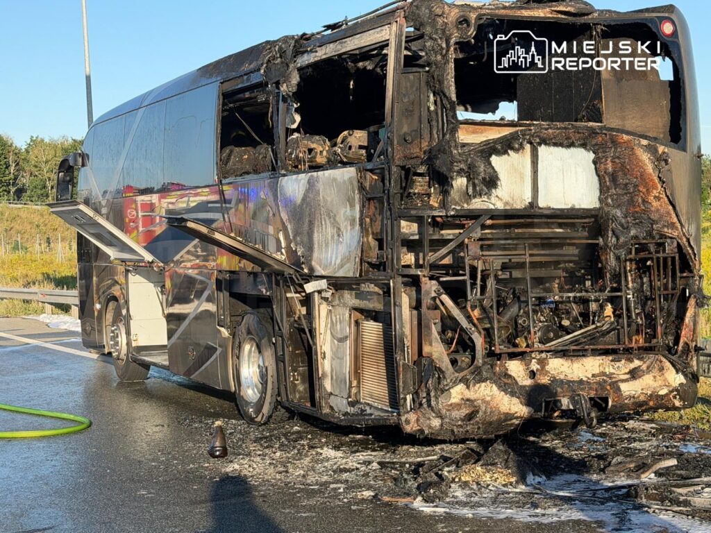 Spalony autobus turystyczny stojący na poboczu drogi, z widocznymi uszkodzeniami i śladami pożaru.