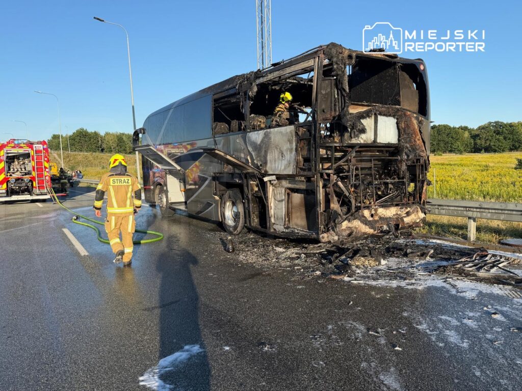 Strażak w żółtym mundurze podchodzi do spalonego autobusu na drodze, obok stoi wóz strażacki.