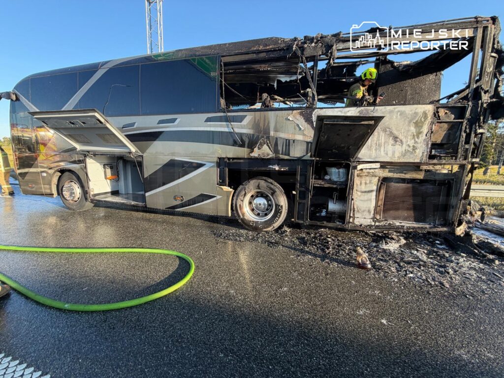Spalony autobus turystyczny z otwartymi drzwiami, przy którym pracuje strażak, na mokrej nawierzchni drogi.