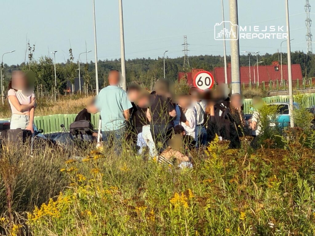 Grupa osób rozmawiająca obok drogi, w tle znak ograniczenia prędkości do 60 km/h oraz zarośla.