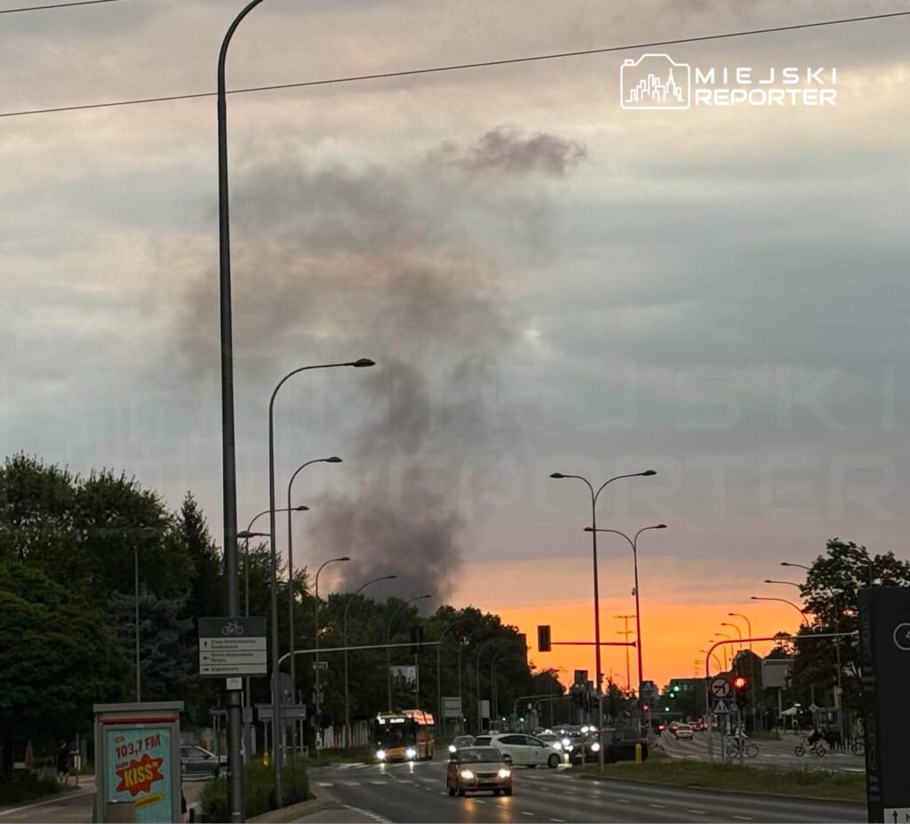 Czarny dym unosi się nad horyzontem w tle, podczas gdy ruch uliczny toczy się wzdłuż drogi w mieście.