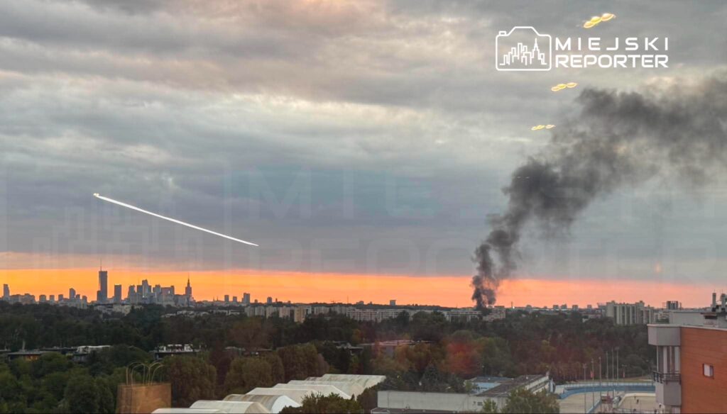 Widok na miasto w tle z zachodzącym słońcem; w powietrzu widoczny dym czarny unoszący się z dolnej części kadru.
