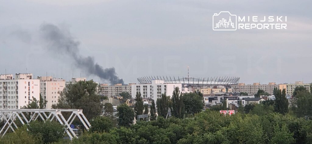 Czarny dym unosi się nad miastem, w tle widoczny stadion oraz blokowiska otoczone zielenią.