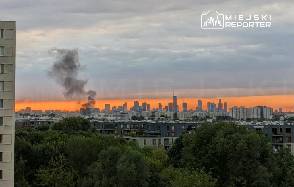 Widok na miejskie skyline z dymem unoszącym się w tle, na tle zachodzącego słońca.