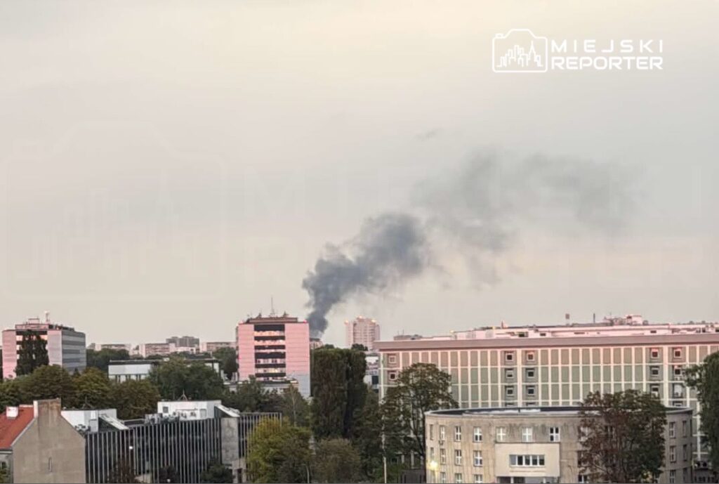 Czarny dym unoszący się z komina w miejskim krajobrazie, w tle nowoczesne budynki mieszkalne.